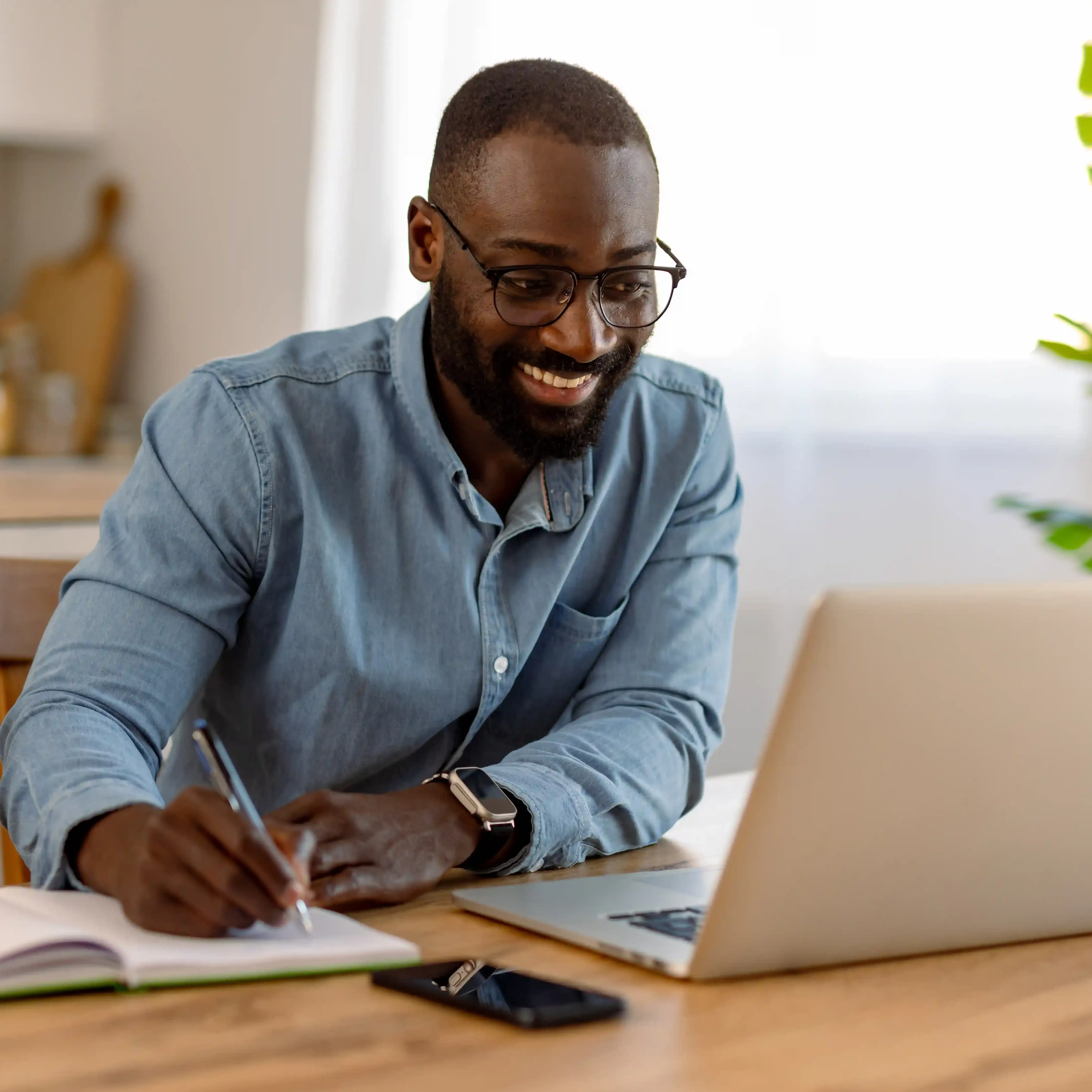 A man wearing glasses and blue button down smiles while writing notes and looking at his laptop