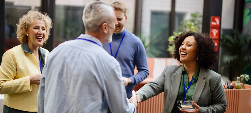 A woman in a suit blazer shakes a man's hand during a conference where they are surrounded by other colleagues