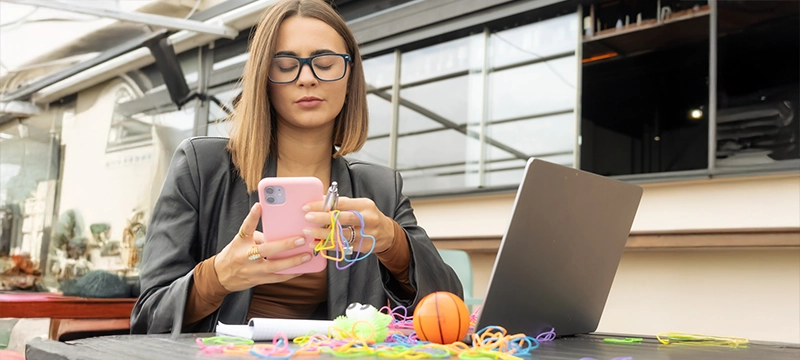 A woman plays with her phone while using her laptop and also playing with fidget toys, symbolizing adult adhd