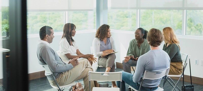A diverse group of adults sit in a circle with a mental health professional, representing a community mental health center