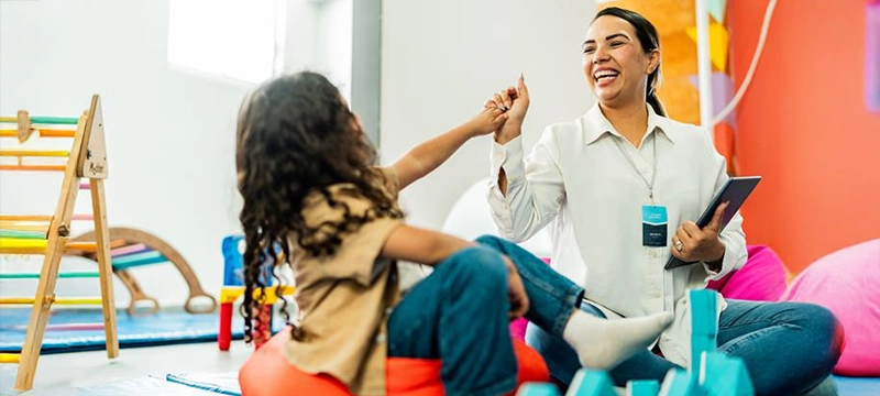 A young girl celebrates with an adult woman holding an iPad in a classroom or playroom setting, representing a happy moment of a psychologist who has administered the RIAS-2 NU