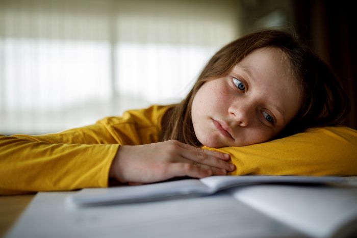 A young girl with blue eyes rests her head on a notebook looking somber