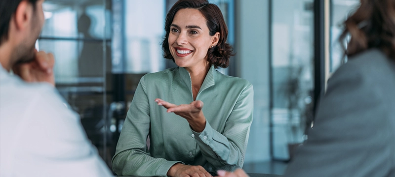 A woman in professional clothing smiles while gesturing in agreement with the man sitting across from her in an office setting