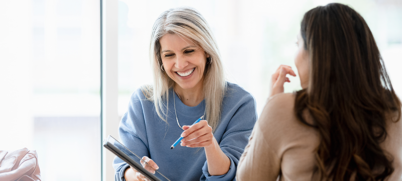 A blonde woman smiles while pointing at a tablet screen while conversing with a brunette woman across from her