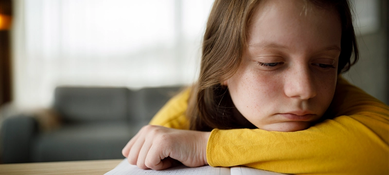 A child in a yellow shirt looks forlorn while resting their head on their arm as they struggle with homework