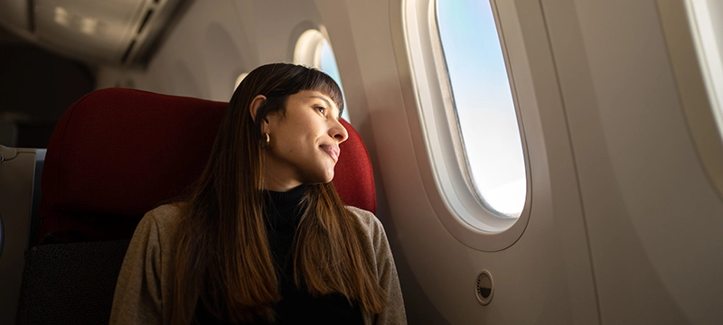 A brunette woman smiles softly while looking out the window of an airplane during a flight, representing travel and the happiness that is linked to traveling.